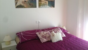 Une chambre à coucher comprenant un lit, des oreillers, une table de chevet, une lampe et deux tableaux représentant des paysages côtiers, accrochés au mur.
