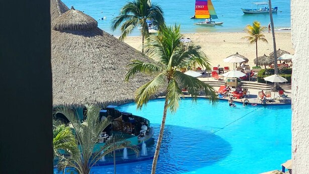 Uitzicht op een resort met een zwembad, palmbomen en een zeilboot op het water.