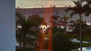 Zonsondergang vanaf een balkon met uitzicht op een strand, palmbomen en een zwembad.