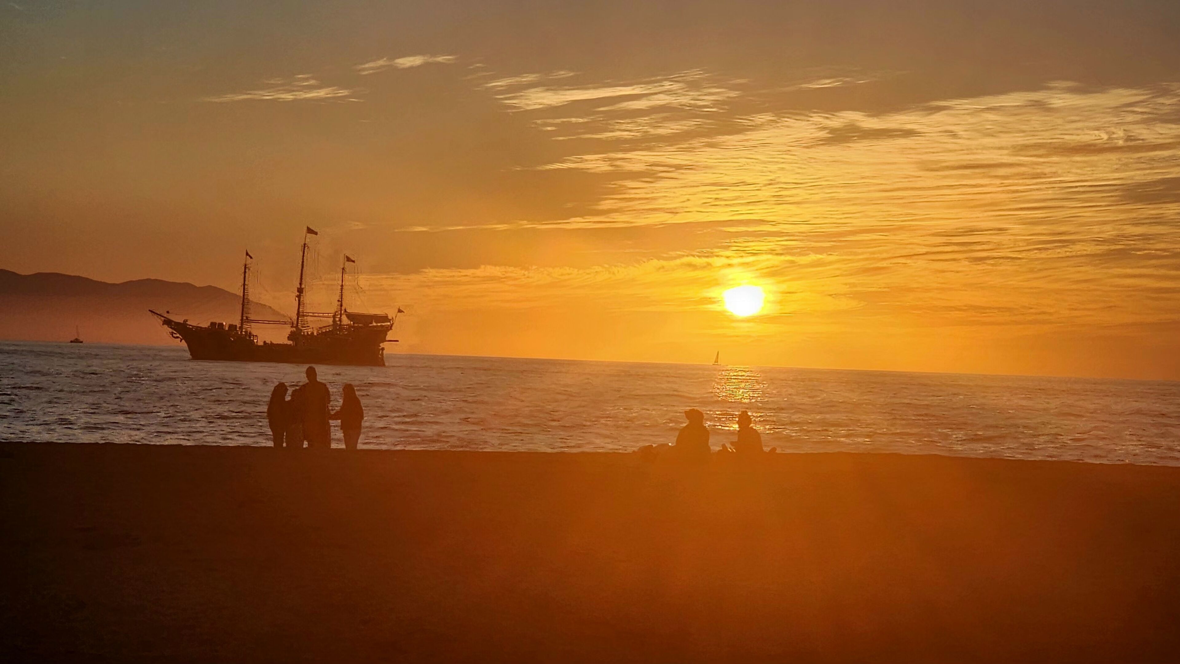 A sunset over the ocean with a ship and people on the shore.