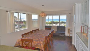 A dining area with a table set for six, large windows with curtains, a view of the ocean, and a kitchen with cabinets and a countertop. - Gathering Place overlooking beach, pier, and downtown Cayucos (Cayucos)