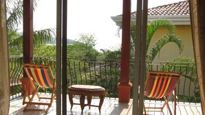 A balcony with two striped chairs and a small round table, overlooking a garden with palm trees. - Fabulous Spacious Villa Only Steps Away from the Beach!  (Playa Hermosa)