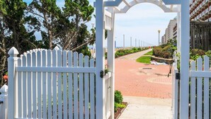 A gated entrance with a white picket fence leading to a paved path. - Virginia Beach Oceanfront Studio, Lawn, Boardwalk, Pool, Beach (Virginia Beach)