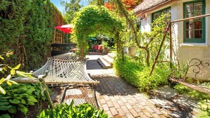 A garden pathway leading to a rustic house with a white exterior and green shutters. There is a hammock and a red umbrella in the garden.