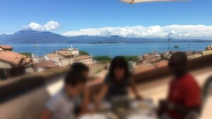 A dining table with plates and glasses, set against a backdrop of a coastal view with buildings and mountains. - Appartment with unique view on the Garda Lake (Desenzano del Garda)
