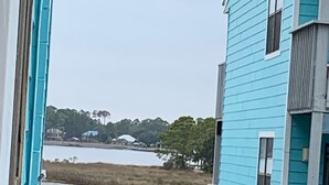 A view from inside a building looking out to a body of water, a grassy area, and a blue building with a balcony.