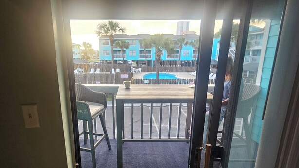 A view from a room to an outdoor pool area with a wooden deck, lounge chairs, and a fenced swimming pool.