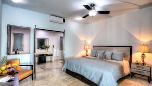 A bedroom with a bed, bedside lamps, a ceiling fan, a bathroom with a mirror, and a dining area with fruit and a glass of wine.