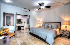 A bedroom with a bed, bedside lamps, a ceiling fan, a bathroom with a mirror, and a dining area with fruit and a wine glass.