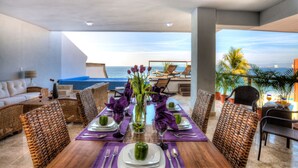 A dining area with a view of the ocean, featuring a wooden table set with purple tablecloths, wicker chairs, and a vase with flowers.