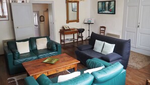 Un salon avec deux canapés verts, une table basse en bois et un canapé bleu foncé. On y trouve une porte, un miroir et un tableau encadré au mur.