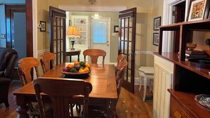 A dining area with a wooden table, chairs, and a fruit bowl. There is a chandelier above the table and a bookshelf with framed pictures to the right.