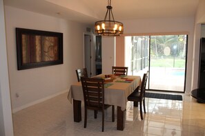 A dining area with a table set for four, chairs, a chandelier, a framed artwork on the wall, and a view of the outdoors through a glass door.