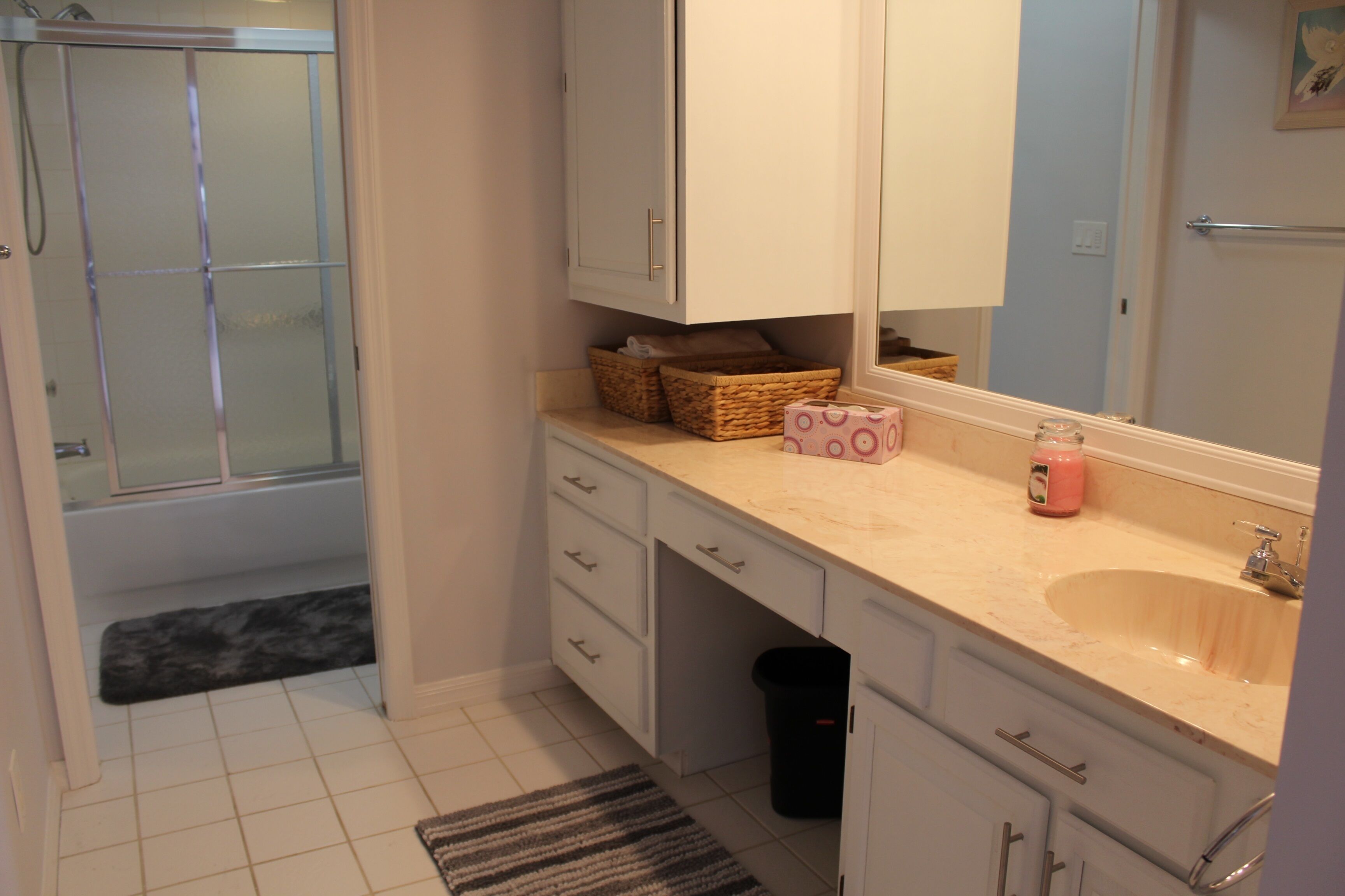 A bathroom with a shower, a sink, and a mirror. There are baskets on the counter and a trash can under the sink.