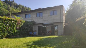 Edificio de dos pisos con un patio amoblado, que incluye una mesa y sillas, rodeado por un césped cuidado y abundante vegetación.