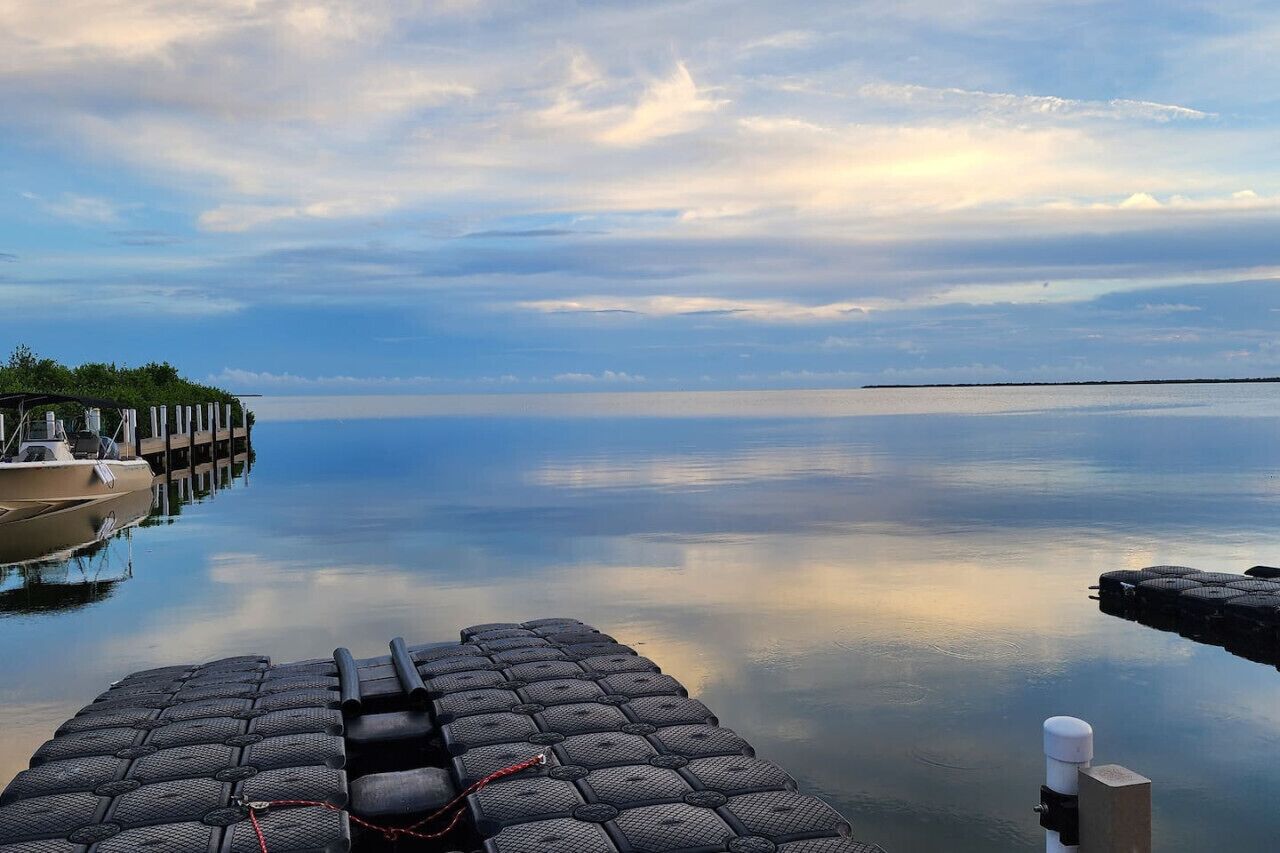 A calm waterfront with a dock extending into the water, a boat moored at the dock, and a row of lounge chairs facing the water.