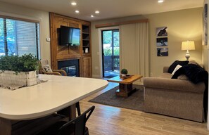 A living room with a fireplace, a television mounted on a wooden wall, a dining area with a table and chairs, and a sofa.