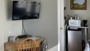 Un espace de vie compact comprenant un téléviseur à écran plat fixé au mur, un bureau en bois avec une chaise, un petit réfrigérateur et un four à micro-ondes posés sur un plan de travail blanc.