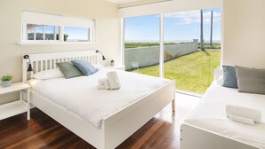 A bedroom with a white bed, a bedside table with a lamp, and a view of a grassy area through large windows. - BOWERBIRD VIEW - Breathtaking Ocean Views (Busselton)