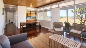 A spacious living area with a wooden floor, a sofa, two wicker chairs, a bench, and a view of the beach through large windows. - BOWERBIRD VIEW - Breathtaking Ocean Views (Busselton)