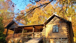 A log cabin with a porch and a stone foundation, surrounded by autumn foliage.