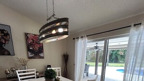 A dining area with a white table, chairs, a hanging light fixture, and a view of a swimming pool through large windows. - Fantastic Lakefront Home With Private Yard and Pool (Fort Lauderdale)