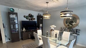 A dining area with a glass table, white chairs, and a patterned rug. There is a television and a cabinet in the background. - Fantastic Lakefront Home With Private Yard and Pool (Fort Lauderdale)