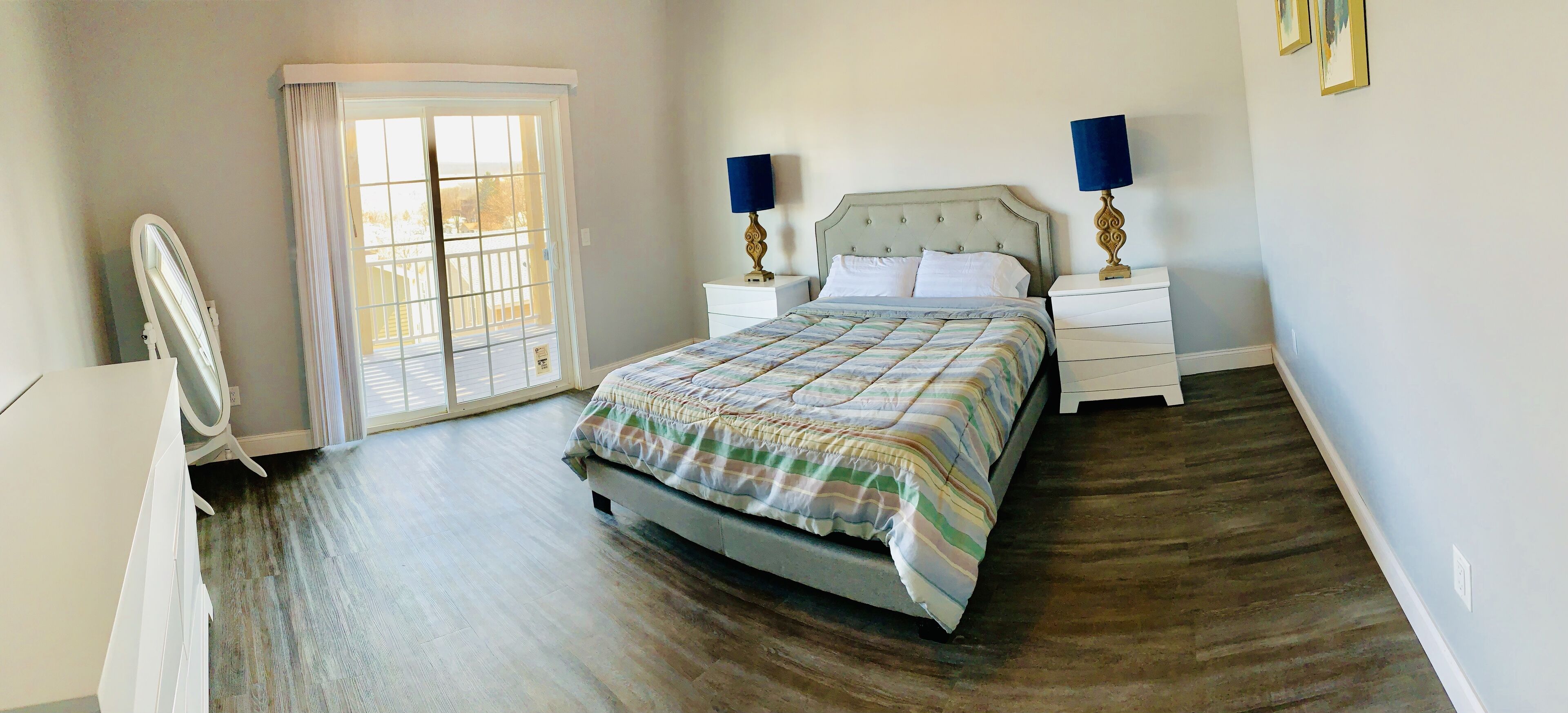 A bedroom with a bed, two bedside tables with lamps, a dresser, a mirror, and a sliding glass door leading to a balcony.