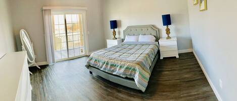 A bedroom with a bed, two bedside tables with lamps, a dresser, a mirror, and a sliding glass door leading to a balcony.