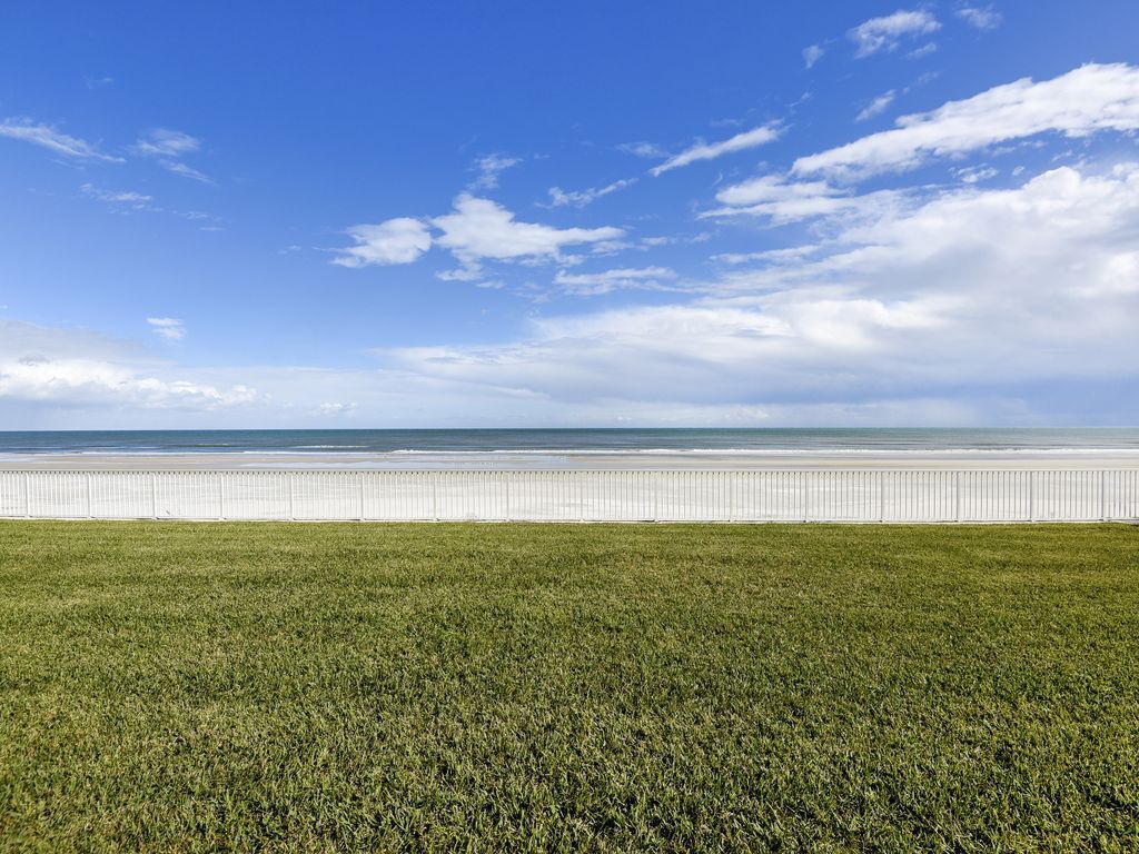 Un campo cubierto de hierba con una cerca blanca y una playa al fondo.