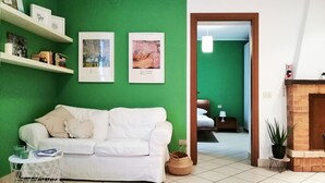 Un salon avec un canapé blanc, des murs verts et une cheminée en bois. Des tableaux encadrés ornent le mur, et une petite table avec un panier se trouve à proximité.