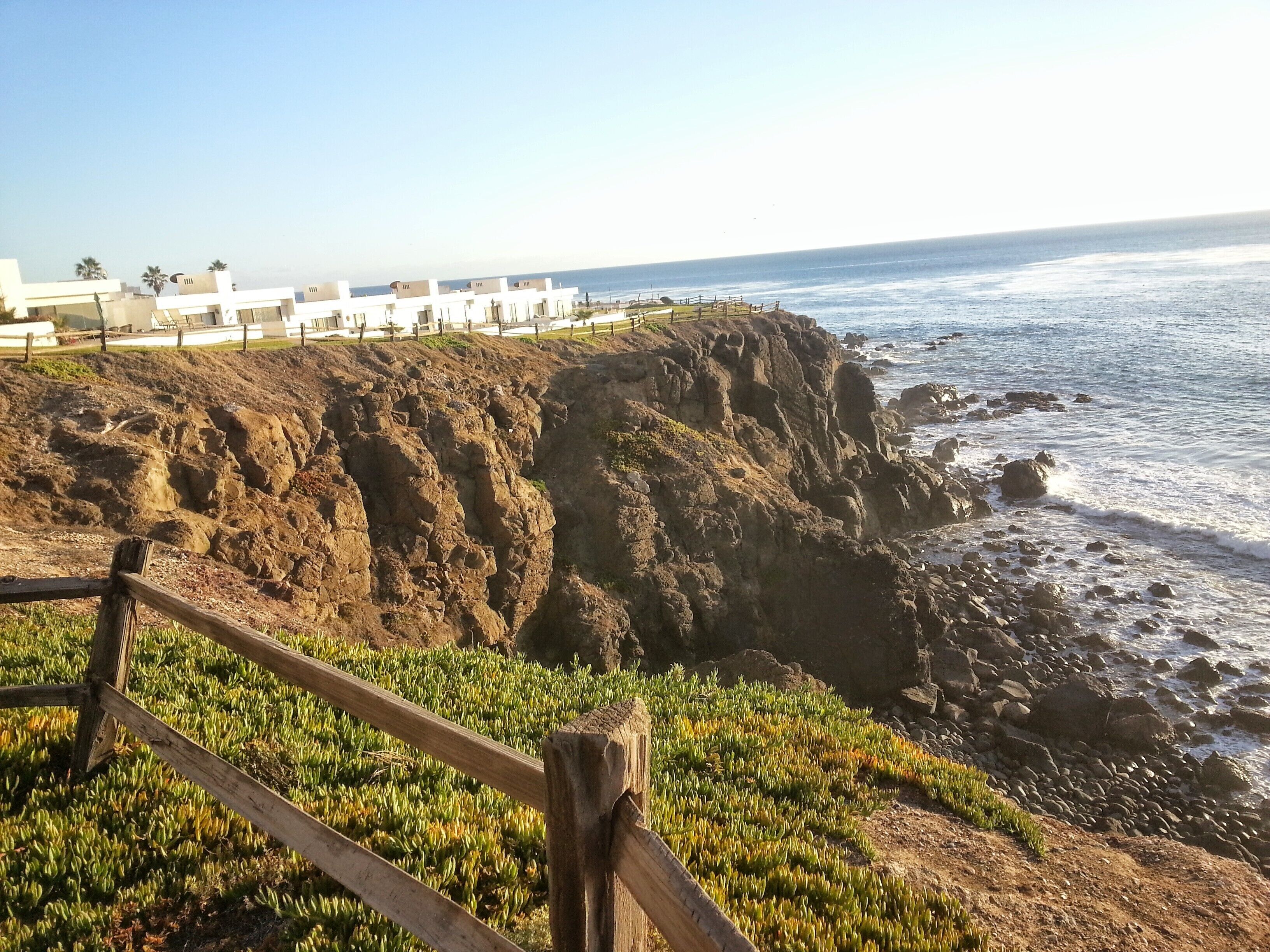A coastal view with rocky cliffs, a wooden fence, and a building with a flat roof in the background.