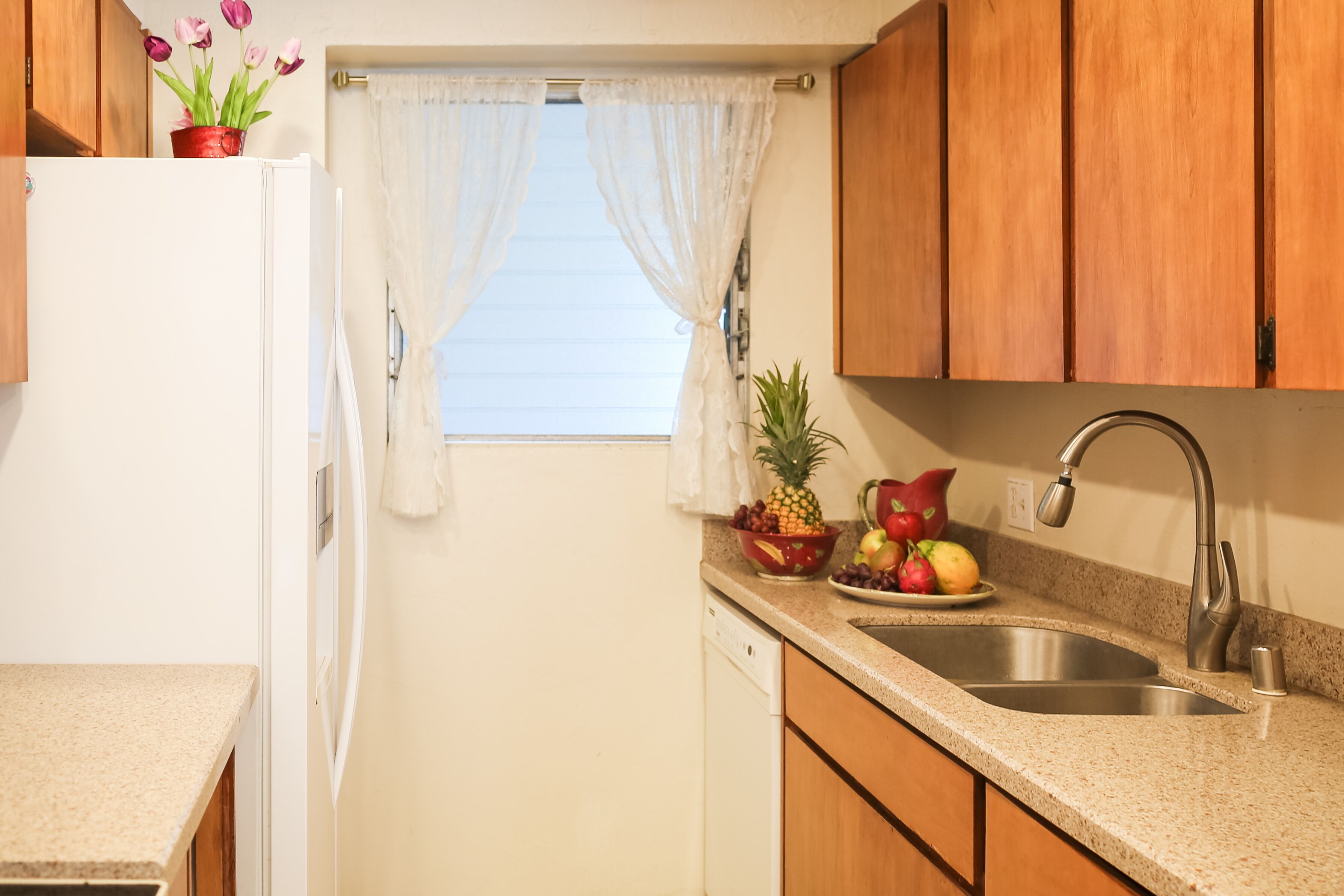 A compact kitchen with wooden cabinets, a white refrigerator, a sink, and a window with sheer curtains.