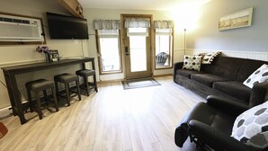 Una sala de estar con un sofá marrón oscuro, un sillón de cuero negro y una mesa de comedor de madera con tres taburetes. Hay un televisor de pantalla plana en la pared, una ventana con vistas y una puerta que da al exterior.
