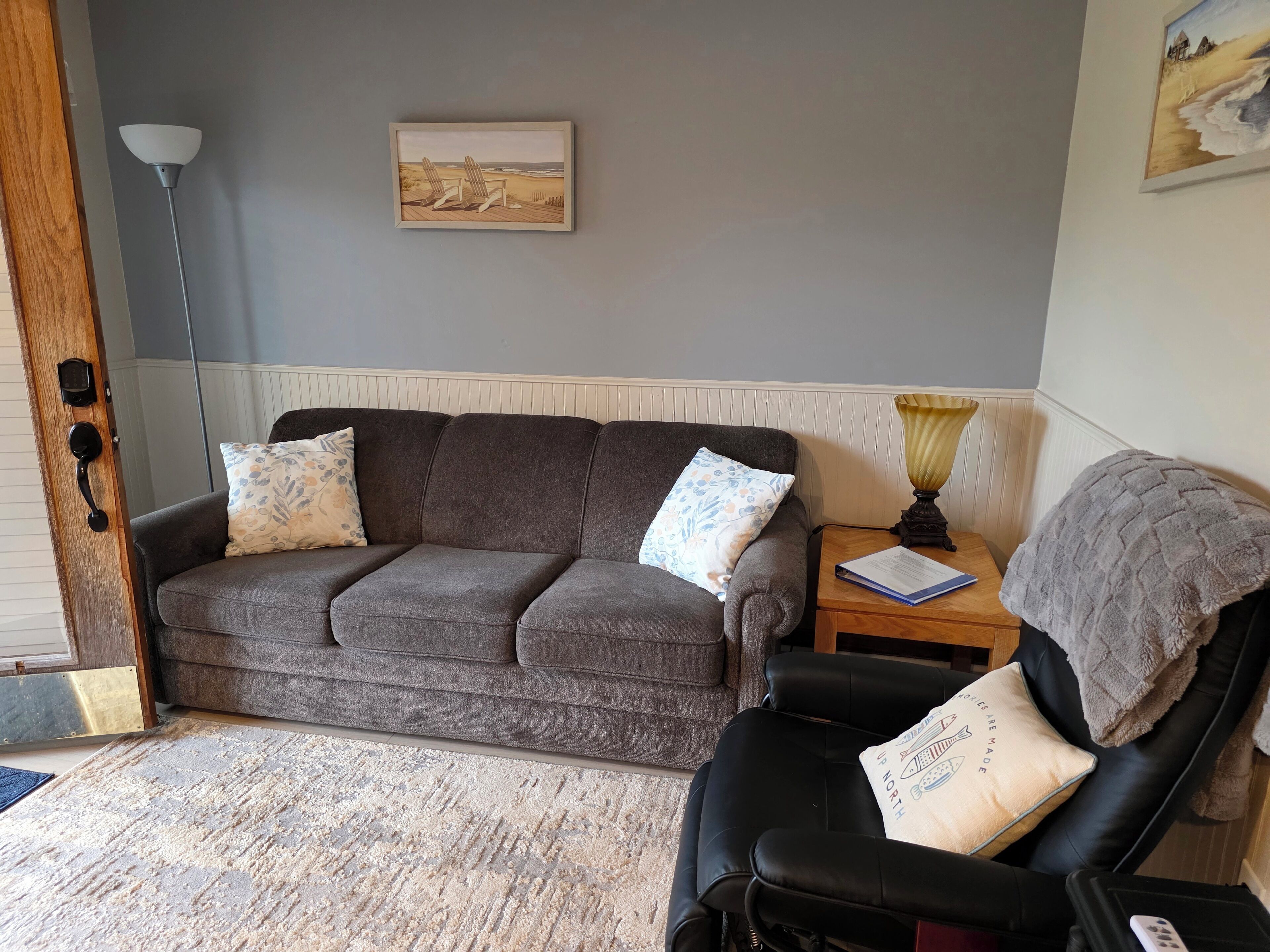 Een woonkamer met een grijze bank, een zwarte fauteuil, een tafeltje met een lamp en een ingelijst schilderij aan de muur.
