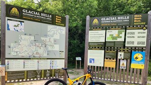 Una bicicleta de montaña amarilla estacionada frente a un letrero de inicio de sendero con mapas e información sobre el Sendero y Área Natural Glacial Hills.