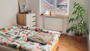 Une chambre à coucher avec un lit, une commode en bois, un radiateur, une fenêtre donnant sur des immeubles et une plante en pot.
