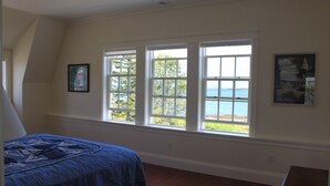 Une chambre avec un grand lit, une vue sur la mer à travers trois fenêtres et un tableau encadré au mur.