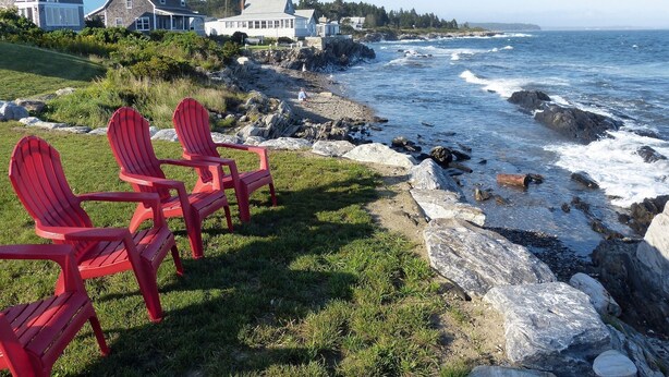 Une vue sur la côte, avec des chaises Adirondack rouges, un rivage rocheux et des maisons en arrière-plan.
