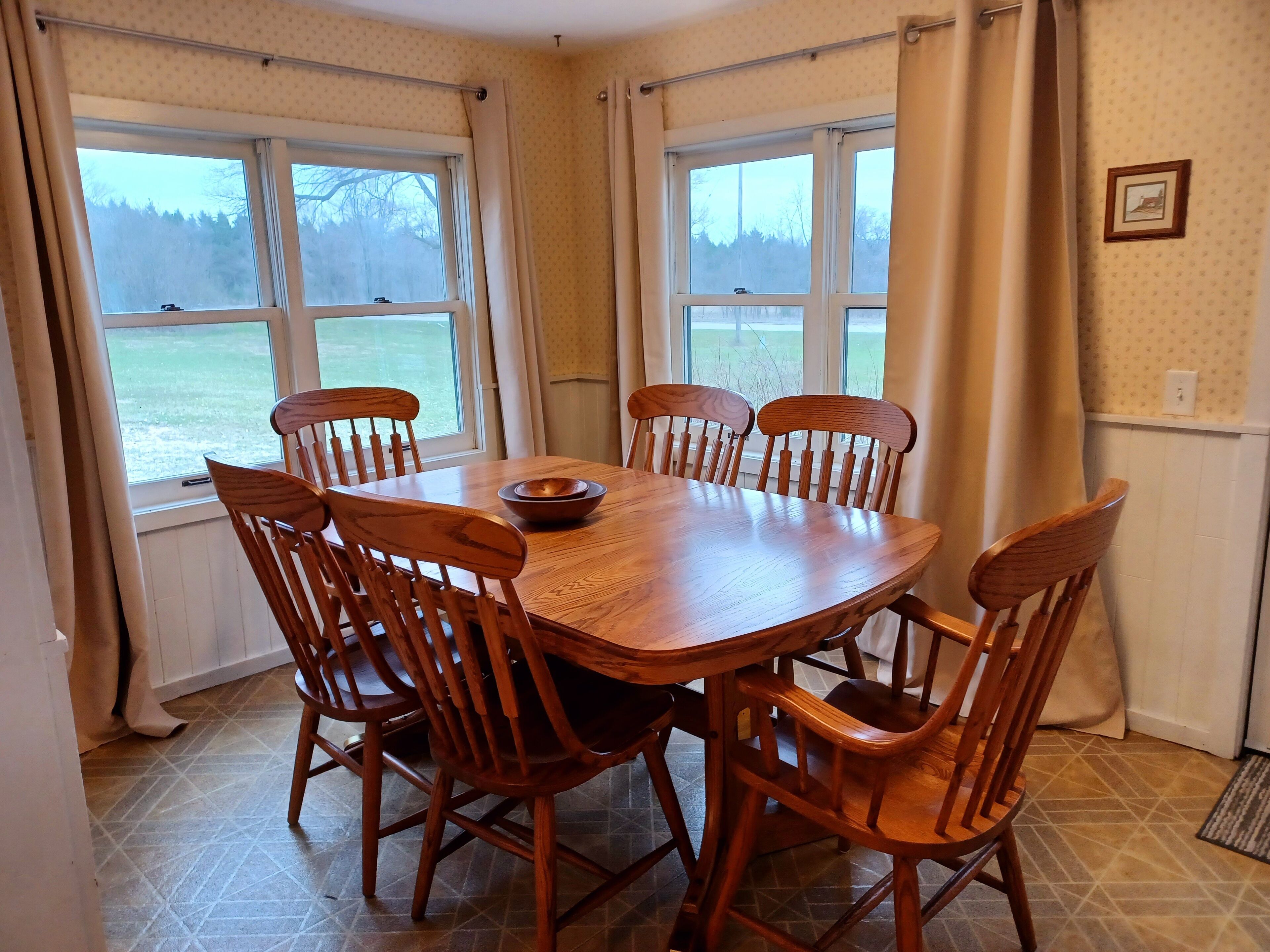 A dining room with a wooden table and chairs, large windows with curtains, and a view of a body of water.