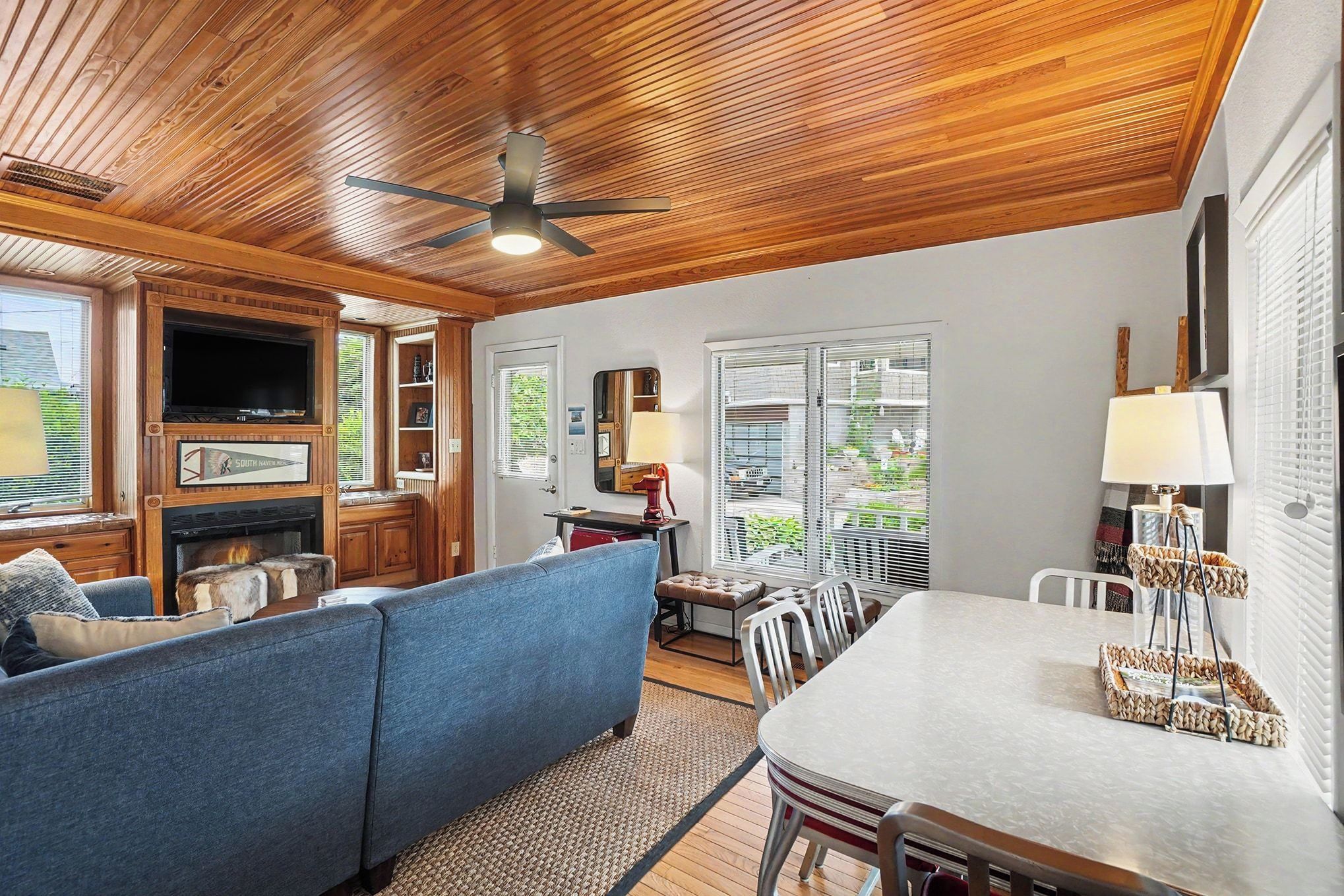 A cozy living room with a wooden ceiling, a television, a fireplace, a blue sofa, and a dining area with a table and chairs.