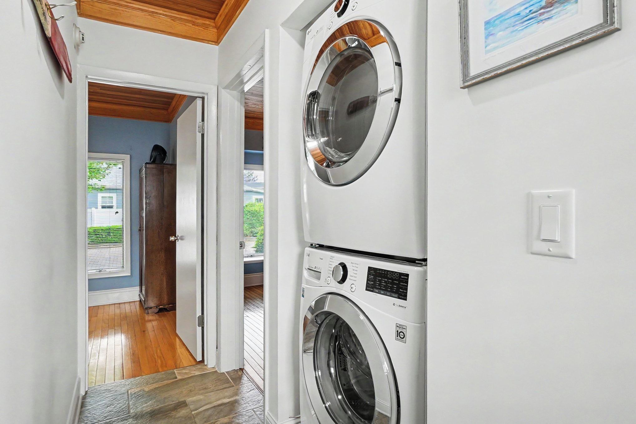A laundry room with two stacked front-loading washing machines, a wooden door leading to a hallway, and a framed picture on the wall.