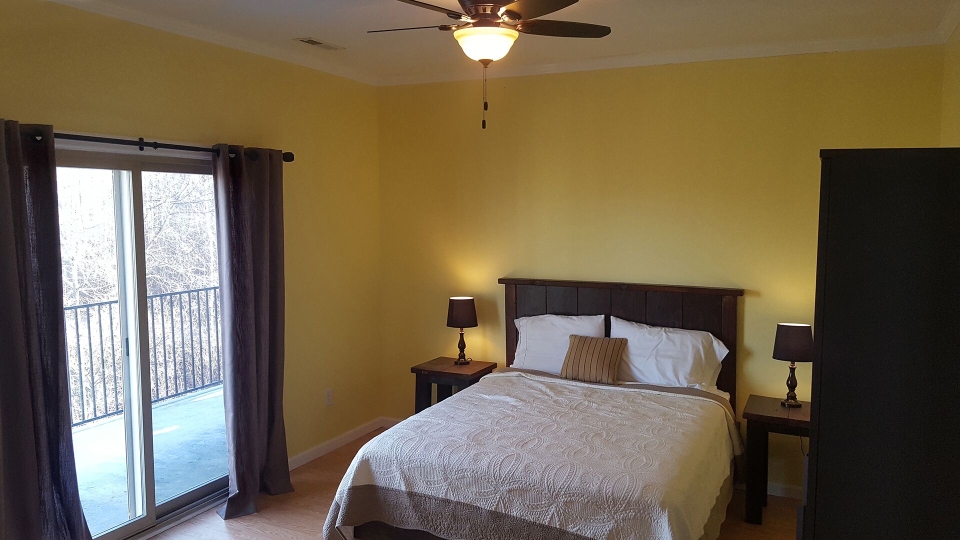 A bedroom with a bed, two bedside tables with lamps, a ceiling fan, and a large window with curtains.