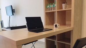 Um laptop sobre uma mesa de madeira com uma cadeira, uma estante com garrafas e uma caneca, e uma TV na parede.