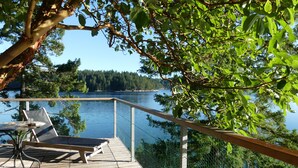 Una terraza de madera con mesa y sillas da a un lago, rodeado de árboles y una valla.