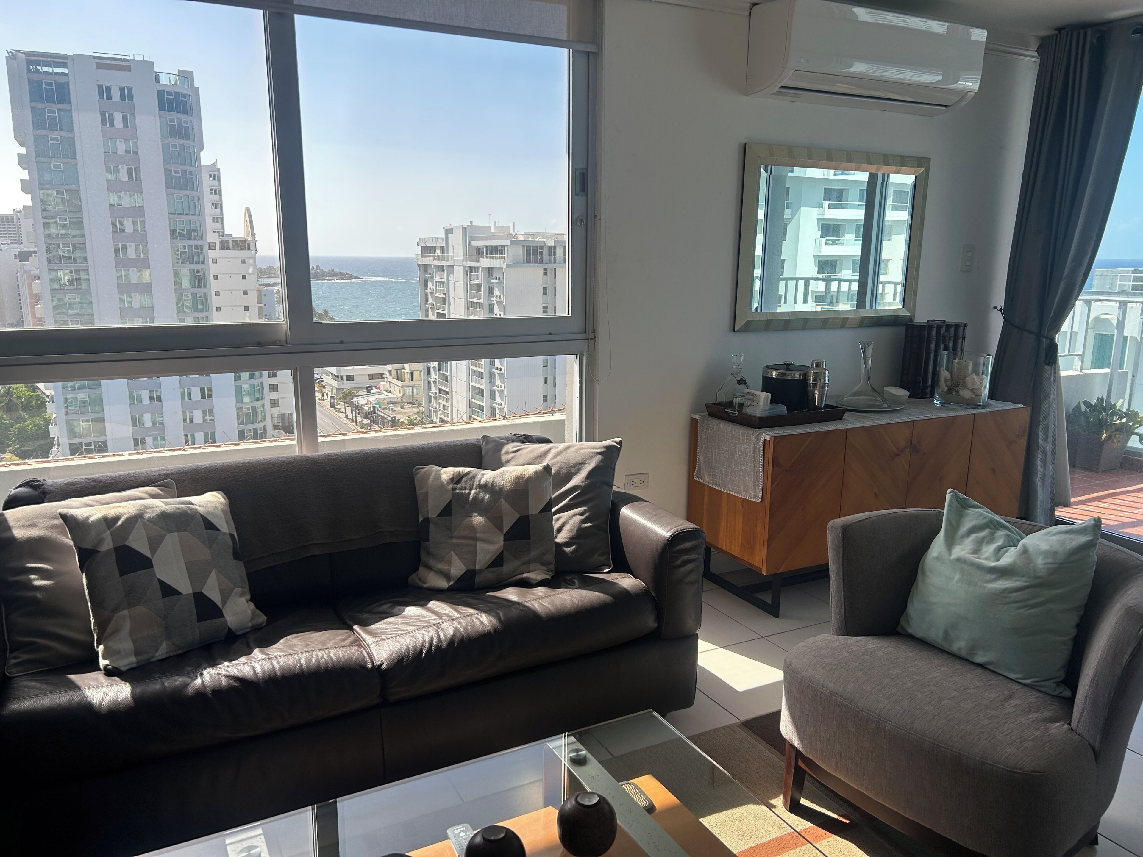 A modern living room with a leather sofa, a glass coffee table, and a view of the ocean and buildings.