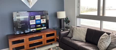 A living room with a flat-screen TV on a wooden stand, a leather sofa, a glass coffee table, and a large window with a view of a body of water.