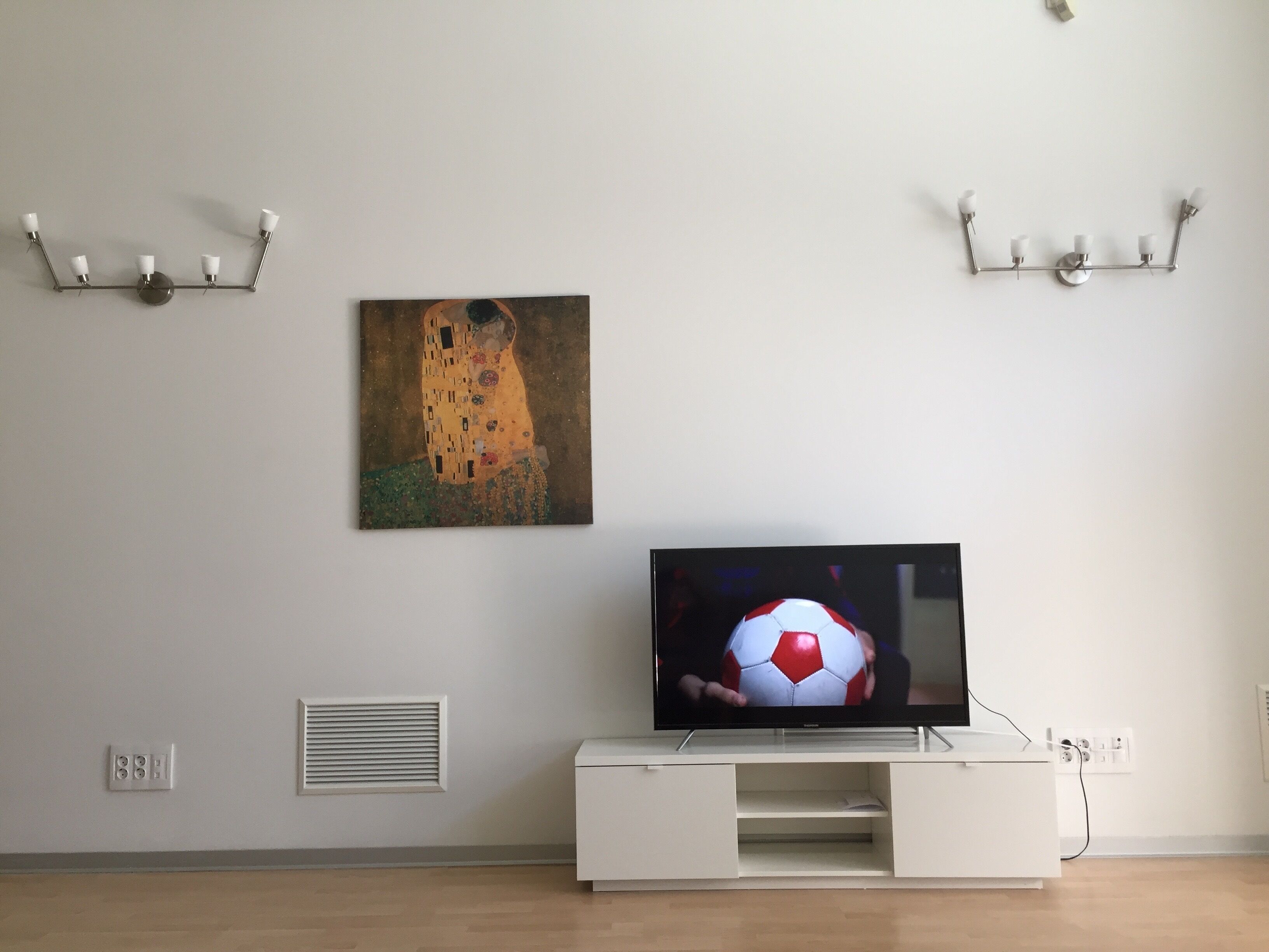 A modern living room with a flat-screen TV on a white entertainment unit, a wall-mounted painting, and a wall-mounted light fixture.