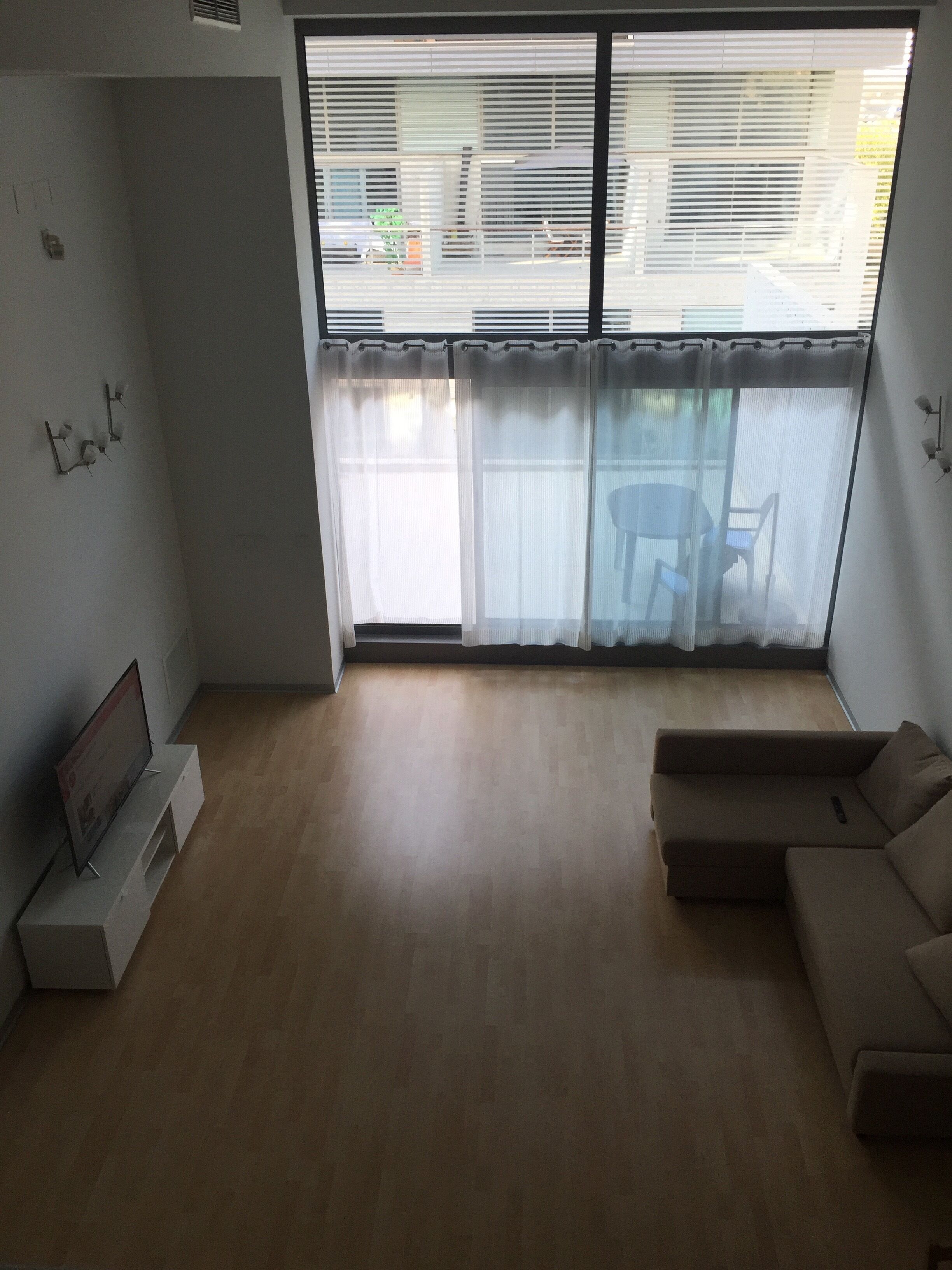 Una sala de estar con un ventanal grande, un sofá, un televisor sobre un mueble bajo y un aplique de pared.