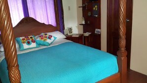 A four-poster bed with a blue bedspread and floral pillows, a wooden nightstand with a lamp, and a wall-mounted shelf with various items.